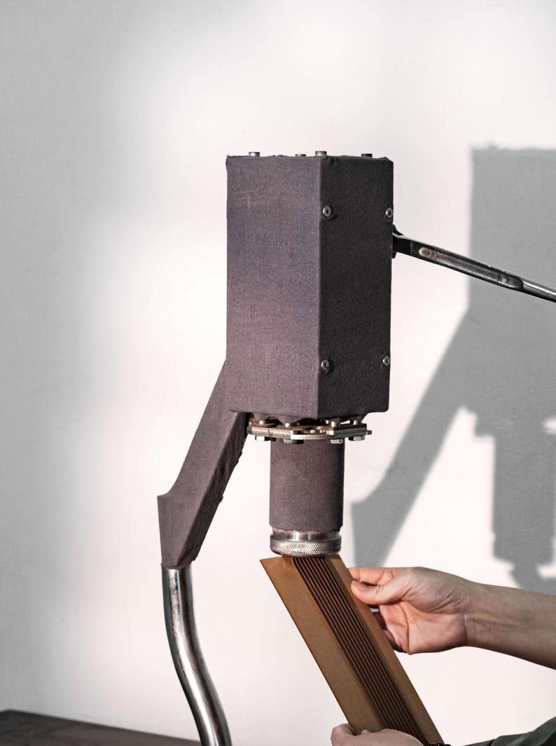Op de afbeelding: Een donkergrijze industri&euml;le machine met een rechthoekige bovenkant en een gebogen metalen arm. Een persoon houdt een houten stuk onder de cilindrische basis van de machine. De machine heeft een complex mechanisch ontwerp.