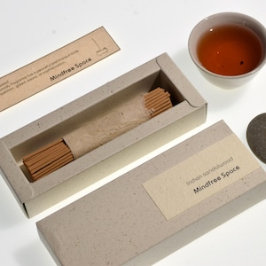 Op de afbeelding: Een open doos met Indiase sandelhout wierookstokjes met de tekst "Mindfree Space" op het etiket. Een klein kopje thee en een stenen wierookhouder zijn ook aanwezig. De verpakking is lichtbeige.