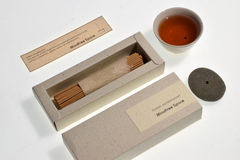 Op de afbeelding: Een open doos met Indiase sandelhout wierookstokjes met de tekst "Mindfree Space" op het etiket. Een klein kopje thee en een stenen wierookhouder zijn ook aanwezig. De verpakking is lichtbeige.