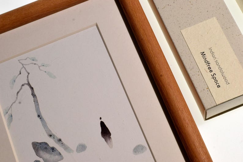 Op de afbeelding: Aquarel schilderij van een tak met bladeren, omlijst in een houten frame. Een doos met de tekst "Indian sandalwood Mindfree Space" staat naast het ingelijste kunstwerk. Het kunstwerk heeft een minimalistische stijl.