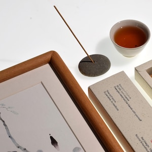 Op de afbeelding: Een compositie met een ingelijst kunstwerk, een kopje thee, wierook en een stenen wierookhouder. Het kunstwerk is een zwart-witte inkttekening. Wierookstokjes zitten in een doos met tekst. De thee zit in een kleine witte keramische beker.
