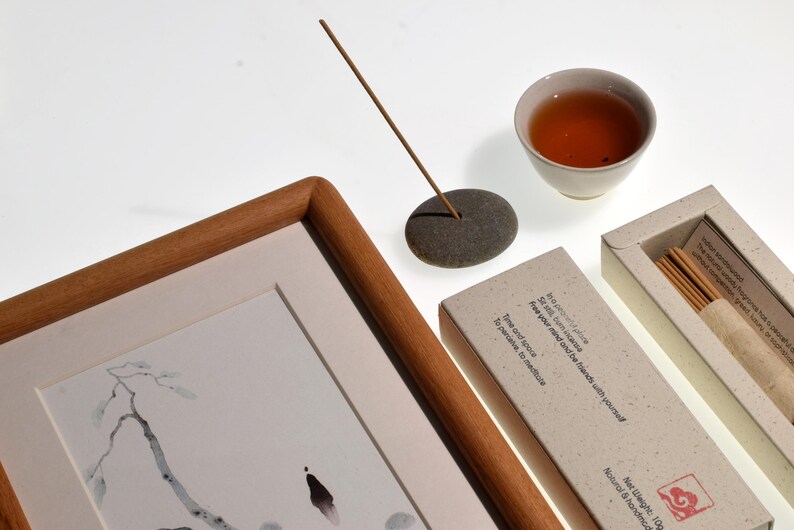 Op de afbeelding: Een compositie met een ingelijst kunstwerk, een kopje thee, wierook en een stenen wierookhouder. Het kunstwerk is een zwart-witte inkttekening. Wierookstokjes zitten in een doos met tekst. De thee zit in een kleine witte keramische beker.