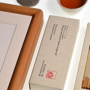Op de afbeelding: Een compositie met een ingelijst kunstwerk, wierookstokjes en een kopje thee. De wierook zit in een natuurlijk gekleurde doos met tekst. Het kunstwerk zit in een houten lijst. De thee zit in een witte keramische beker.
