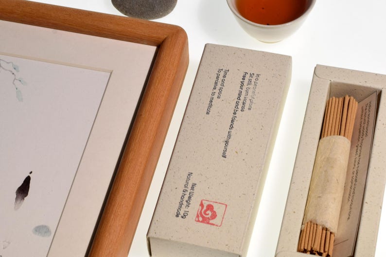 Op de afbeelding: Een compositie met een ingelijst kunstwerk, wierookstokjes en een kopje thee. De wierook zit in een natuurlijk gekleurde doos met tekst. Het kunstwerk zit in een houten lijst. De thee zit in een witte keramische beker.