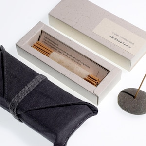 Op de afbeelding: Een set Indiase sandelhout wierookstokjes met een grijze stenen houder, een donkergrijze stoffen hoes en een lichtgrijze doos. De doos is voorzien van het label "Mindfree Space". De wierookstokjes zitten in een lichtgrijze doos met tekst.