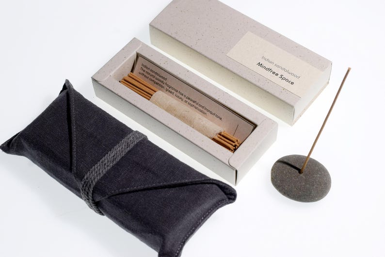 Op de afbeelding: Een set Indiase sandelhout wierookstokjes met een grijze stenen houder, een donkergrijze stoffen hoes en een lichtgrijze doos. De doos is voorzien van het label "Mindfree Space". De wierookstokjes zitten in een lichtgrijze doos met tekst.