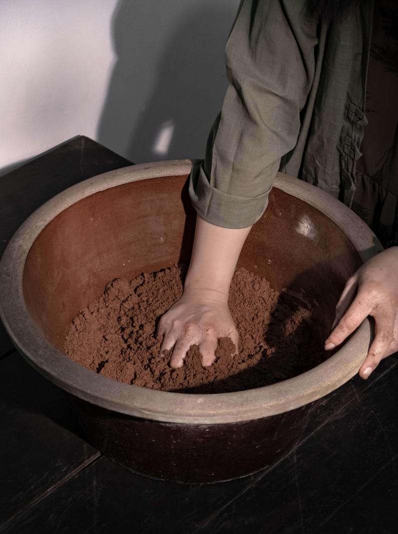 Op de afbeelding: Een grote, bruine keramische kom gevuld met donkerbruin materiaal. De handen van een persoon mengen de inhoud. De kom heeft een brede rand en staat op een donkere houten ondergrond. De persoon draagt een groen overhemd.
