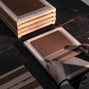 Op de afbeelding: Een houten frame met bruin, getextureerd materiaal. Verschillende frames zijn opgestapeld en een persoon werkt met een ander frame en rangschikt het bruine materiaal. De sc&egrave;ne speelt zich af op een donker houten oppervlak.