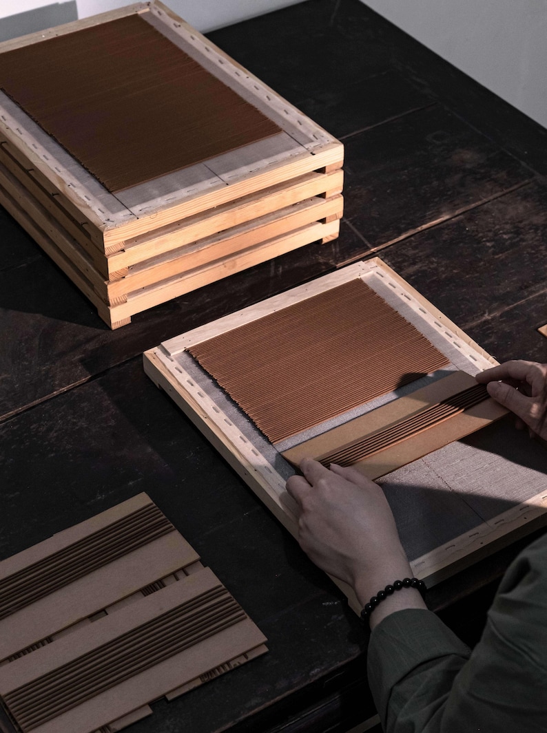 Op de afbeelding: Een houten frame met bruin, getextureerd materiaal. Verschillende frames zijn opgestapeld en een persoon werkt met een ander frame en rangschikt het bruine materiaal. De sc&egrave;ne speelt zich af op een donker houten oppervlak.