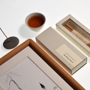 Op de afbeelding: Een compositie met een ingelijst kunstwerk, een kleine kom thee en een doos wierookstokjes. Het kunstwerk heeft een houten lijst en een minimalistische inkttekening. De wierookdoos is gelabeld "Indian sandalwood Mindfree Space."