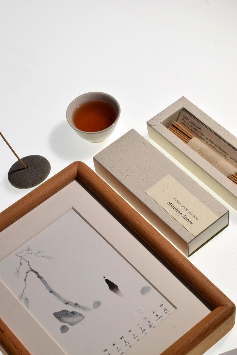 Op de afbeelding: Een compositie met een ingelijst kunstwerk, een kleine kom thee en een doos wierookstokjes. Het kunstwerk heeft een houten lijst en een minimalistische inkttekening. De wierookdoos is gelabeld "Indian sandalwood Mindfree Space."