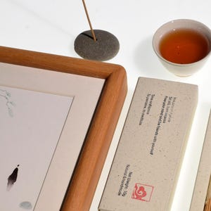 Op de afbeelding: Een zen-stilleven met een ingelijste aquarel, een kopje thee, wierook en wierookstokjes. De schildering toont een minimalistisch landschap. De wierook is aangestoken en de thee is amberkleurig. Op de wierookdoos staat "Bevrijd je geest en wees bevriend met jezelf."