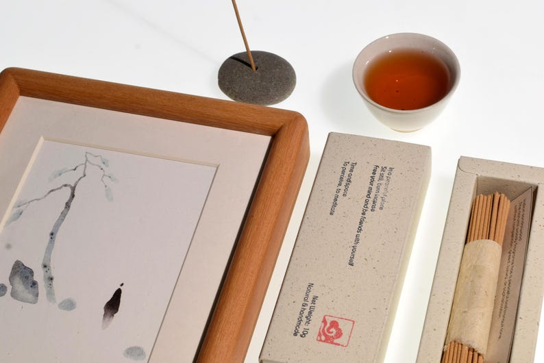 Op de afbeelding: Een zen-stilleven met een ingelijste aquarel, een kopje thee, wierook en wierookstokjes. De schildering toont een minimalistisch landschap. De wierook is aangestoken en de thee is amberkleurig. Op de wierookdoos staat "Bevrijd je geest en wees bevriend met jezelf."