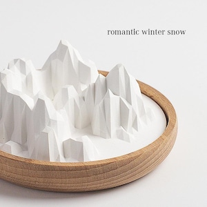 May include: A white, geometric mountain range sculpture inside a round wooden bowl. The text "romantic winter snow" is above the sculpture. The sculpture has a textured, angular design, evoking a winter landscape.