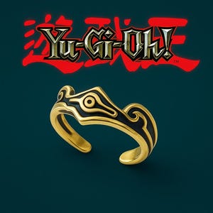 May include: Gold-toned ring with black enamel detailing, featuring a stylized design. The ring is open at the bottom and set against a dark teal background. The Yu-Gi-Oh! logo is visible in the background.