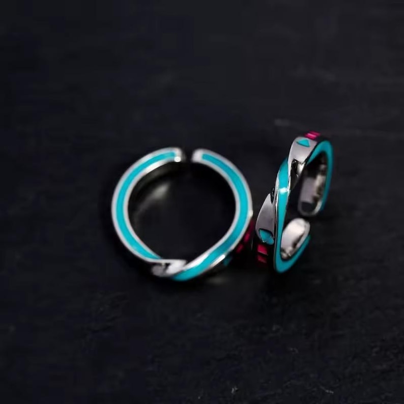 May include: Two silver-toned rings with a twisted design. Each ring features a bright turquoise enamel band and small pink accents. The rings are open-ended, allowing for adjustable sizing. The background is a dark, solid color.