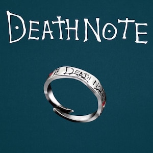 May include: A silver ring with the words "DEATH NOTE" in white script above it. The ring has the words "DEATH NOTE" engraved around the band with red accents. The ring is on a dark teal background.