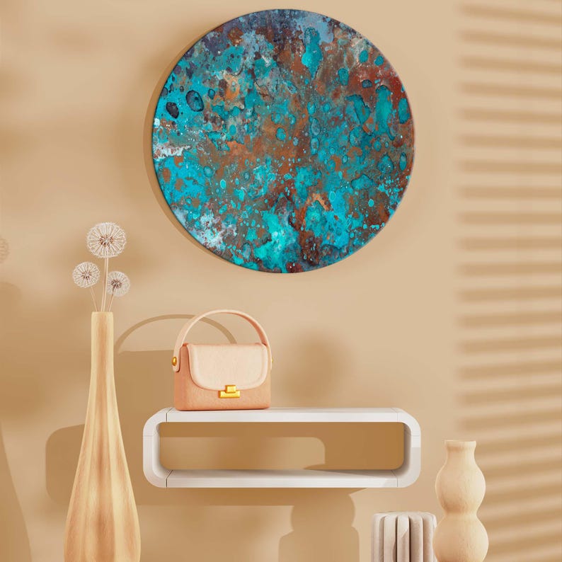 Op de afbeelding: Een rond kunstwerk voor aan de muur met een textuur, abstract ontwerp in tinten van teal, bruin en blauw hangt boven een witte plank. Een kleine, lichtroze handtas met een gouden sluiting staat op de plank, naast een hoge vaas en decoratieve objecten.