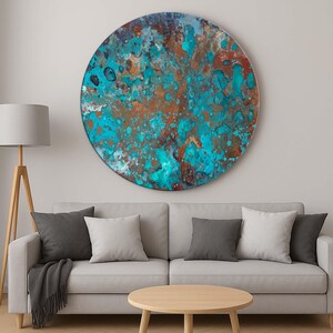 Op de afbeelding: Een groot, rond kunstwerk voor aan de muur met een textuur. Het kunstwerk heeft een mix van turquoise, koper en bruine tinten, waardoor een abstracte, verweerde uitstraling ontstaat. Het stuk is gemonteerd boven een grijze bank met kussens.