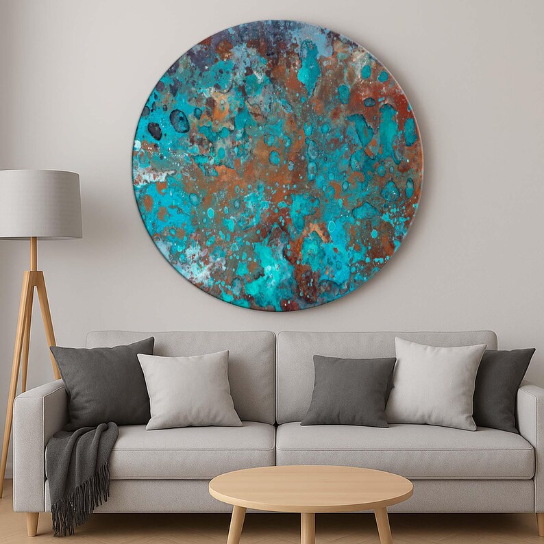 Op de afbeelding: Een groot, rond kunstwerk voor aan de muur met een textuur. Het kunstwerk heeft een mix van turquoise, koper en bruine tinten, waardoor een abstracte, verweerde uitstraling ontstaat. Het stuk is gemonteerd boven een grijze bank met kussens.