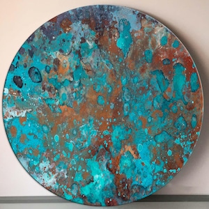 Op de afbeelding: Een rond, abstract kunstwerk met een getextureerd oppervlak. Het kleurenpalet omvat tinten van turquoise, koper en bruin, wat een unieke, verweerde uitstraling cre&euml;ert. Het ontwerp heeft een gevlekt patroon met kleurspatten.