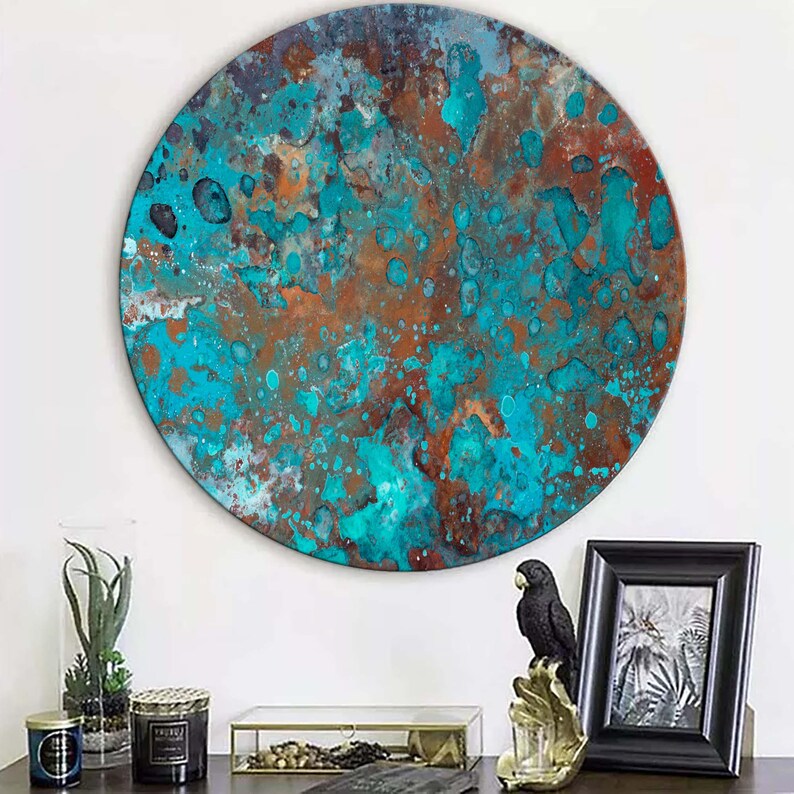 Op de afbeelding: Een groot, rond kunstwerk voor aan de muur met een textuur. Het kunstwerk toont een mix van turquoise, bruine en koperkleuren, wat een abstracte, verweerde uitstraling geeft. Het is aan een witte muur bevestigd, met decoratieve items op een donkere ondergrond.