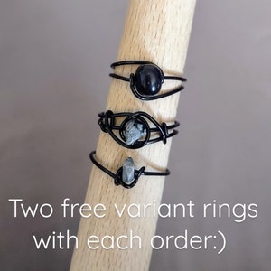May include: Three black wire rings with different stones. The top ring has a black stone, the middle ring has a gray and black stone, and the bottom ring has a gray stone. The rings are displayed on a wooden surface. Two free variant rings with each order.