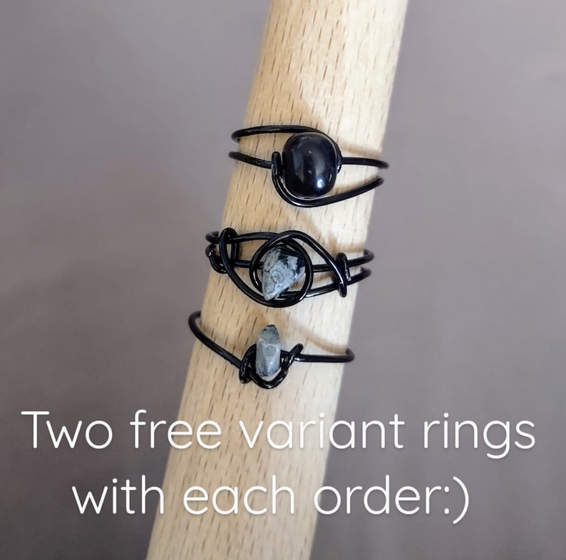 May include: Three black wire rings with different stones. The top ring has a black stone, the middle ring has a gray and black stone, and the bottom ring has a gray stone. The rings are displayed on a wooden surface. Two free variant rings with each order.