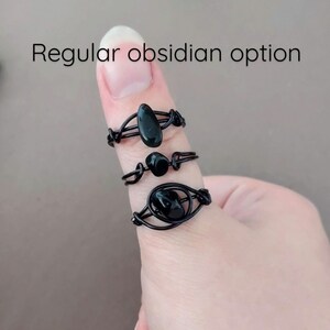 May include: Three black obsidian rings on a finger. The rings are made of wire and have different designs. The top ring has a long, oval-shaped obsidian stone. The middle ring has a round obsidian stone. The bottom ring has a larger, oval-shaped obsidian stone.