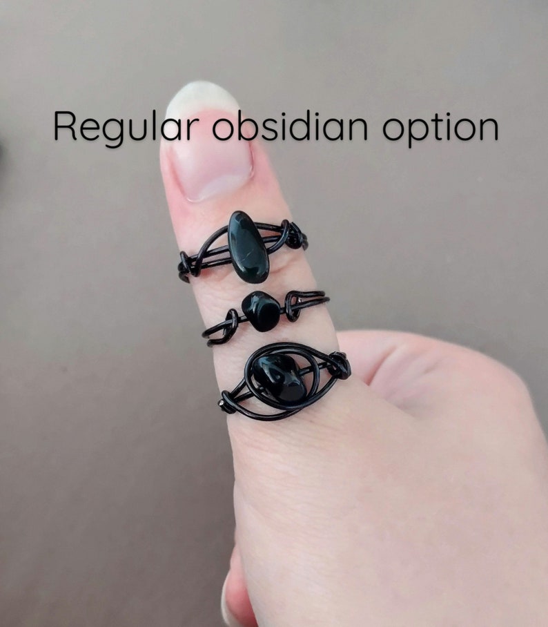 May include: Three black obsidian rings on a finger. The rings are made of wire and have different designs. The top ring has a long, oval-shaped obsidian stone. The middle ring has a round obsidian stone. The bottom ring has a larger, oval-shaped obsidian stone.