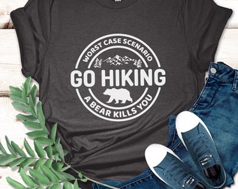 May include: Dark grey heather t-shirt with the text "Worst Case Scenario Go Hiking A Bear Kills You" in a white circular design. The shirt is paired with blue jeans and navy blue sneakers.