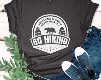 May include: Dark grey heather t-shirt with a white graphic that reads "Worst Case Scenario A Bear Kills You Go Hiking." The shirt is paired with blue jeans and navy blue sneakers. A sprig of green leaves is on the left side of the image.