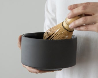 May include: A person holding a dark gray ceramic matcha bowl with a bamboo whisk resting on the rim.