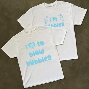 Könnte beinhalten: Zwei weiße T-Shirts mit blauem Text und Blasengrafiken. Auf einem Shirt steht "I ♡ to blow bubbles" und auf dem anderen "I'm bubbles". Die Shirts sind kurzärmlig und haben einen Rundhalsausschnitt.