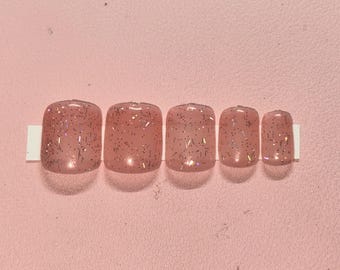 Peut inclure: Un ensemble de cinq faux ongles de tailles variables, disposés sur une bande blanche. Les ongles sont d'une couleur rose translucide et sont recouverts de paillettes argentées. Le fond est rose uni.