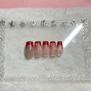 Può includere: Un set di cinque unghie finte con un design a manicure francese. Ogni unghia presenta una base trasparente, una punta rossa e un piccolo fiocco bianco. Le unghie sono disposte su una superficie bianca e soffice all'interno di una cornice decorativa.