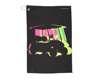 May include: Black golf towel with a colorful golf cart graphic. The golf cart is depicted in shades of pink, yellow, and green, with a black outline. The towel has a metal grommet for hanging.