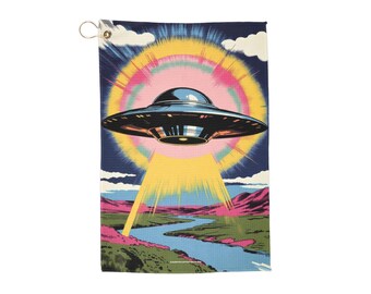 May include: A rectangular golf towel featuring a retro-style illustration of a metallic UFO beaming a yellow light onto a landscape with a river and mountains. The background has a colorful sunburst design.