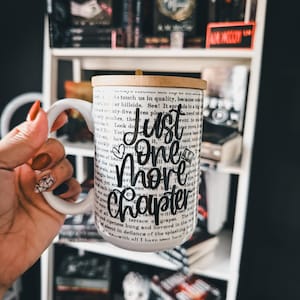 May include: White ceramic mug with the text "Just One More Chapter" in black script. The mug has a wooden lid and is surrounded by a background of books on a shelf.