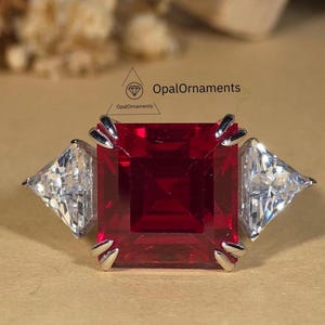 May include: A silver ring featuring a large, square, deep red gemstone flanked by two smaller, triangular, clear gemstones. The ring has a classic design with a focus on the vibrant central stone. The OpalOrnaments logo is visible in the background.
