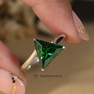 Könnte beinhalten: Ein silberner Ring mit einem dreieckigen grünen Edelstein. Der Ring befindet sich an einem Finger mit goldfarbenem Glitzer-Nagellack.