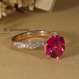 May include: A rose gold ring with a large, oval-cut, ruby-red gemstone. The band features a twisted design with small, clear accent stones. The ring is set against a neutral background with dried flowers and a wooden easel.
