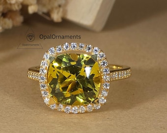 May include: A gold ring with a large, square-cut, yellow-green gemstone. The gemstone is surrounded by a halo of smaller, clear stones. The band of the ring is also adorned with small, clear stones. The ring is set against a neutral background.