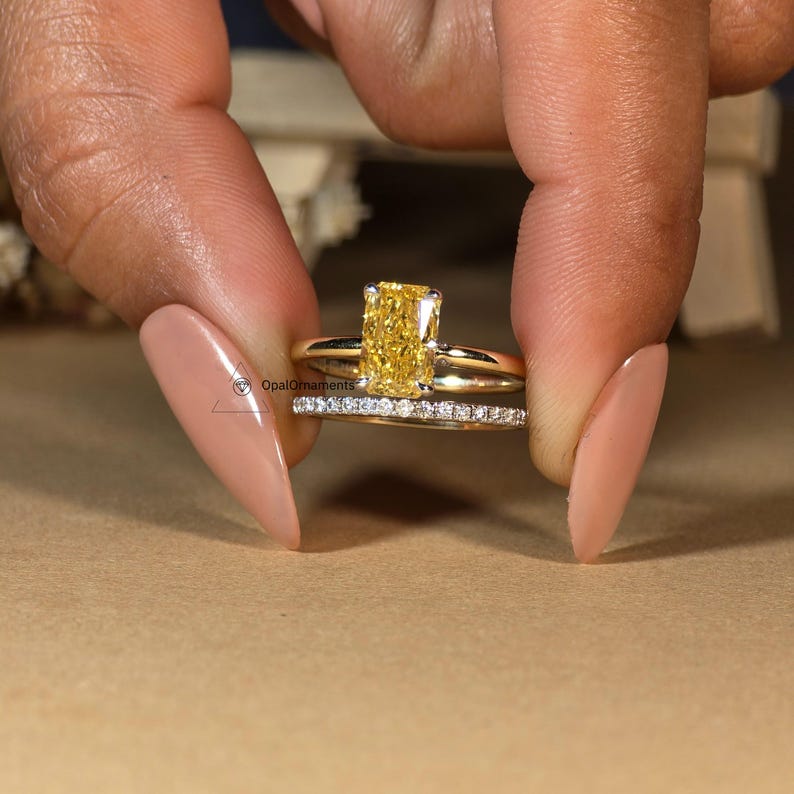 May include: A yellow gold engagement ring with a large, rectangular, yellow diamond. The ring is set with smaller diamonds on the band. The ring is worn on a finger with a light brown nail polish.