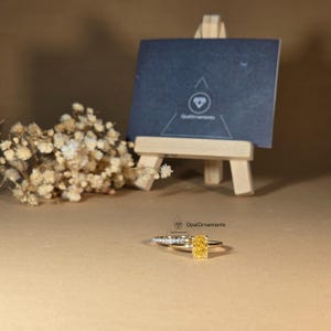 May include: A yellow gold engagement ring set with a large, square-shaped, yellow gemstone. The ring is set with smaller diamonds on the band. The ring is on a brown surface with a small wooden easel and a business card with the text "OpalOrnaments" in the background.
