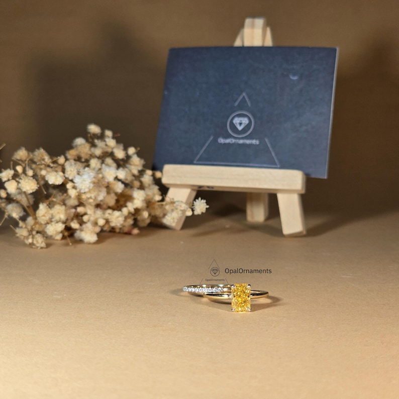 May include: A yellow gold engagement ring set with a large, square-shaped, yellow gemstone. The ring is set with smaller diamonds on the band. The ring is on a brown surface with a small wooden easel and a business card with the text "OpalOrnaments" in the background.