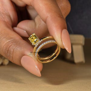 May include: A yellow gold engagement ring set with a square-shaped yellow gemstone surrounded by smaller diamonds. The ring is paired with a matching yellow gold band with a row of diamonds.