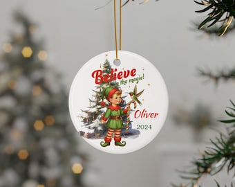 May include: A white ceramic Christmas ornament with a red and green elf holding a candy cane and a hummingbird. The ornament says "Believe in the magic!" and "Oliver 2024".