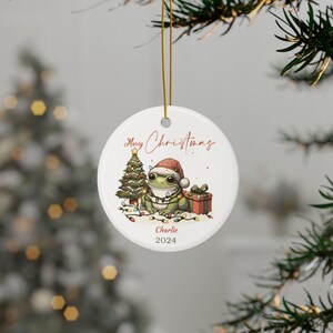 May include: A white ceramic Christmas ornament with a green frog wearing a Santa hat and surrounded by Christmas lights. The ornament also features a small Christmas tree and a red gift box. The text "Merry Christmas" is written in red cursive at the top of the ornament. The text "Charlie 2024" is written in black at the bottom of the ornament.