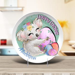 May include: A white ceramic plate with a green background featuring a cartoon koala holding a pink and white Easter egg. The koala is wearing a flower crown and the plate says "Happy Easter" and "Madison".